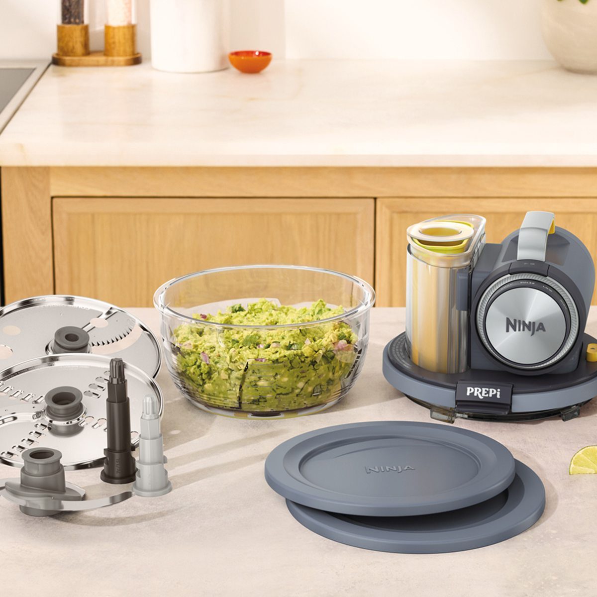 Ninja PREPi kit on kitchen counter showing motor base, bowl of chopped greens, stacked storage lids with Ninja logo, stainless discs, and chop blade accessories